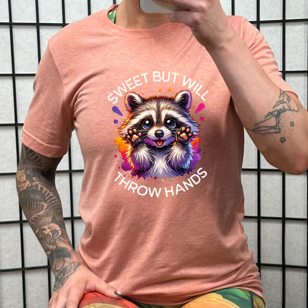 coral shirt with the text "Sweet But Will Throw Hands" and a graphic of a raccoon on it